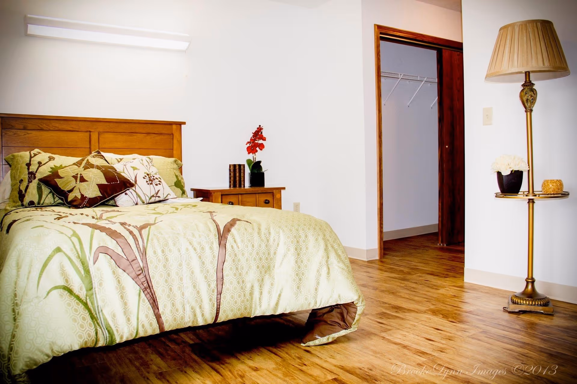 Well-lit bedroom with a wooden bed dressed in floral bedding, a nightstand with a plant, an open closet doorway, and a floor lamp on hardwood floors.