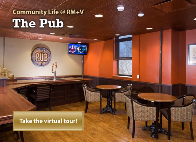 Interior view of a pub-style common area with wooden floors, round tables with patterned chairs, a bar counter with taps, a wall-mounted TV, and warm orange and beige walls with windows.