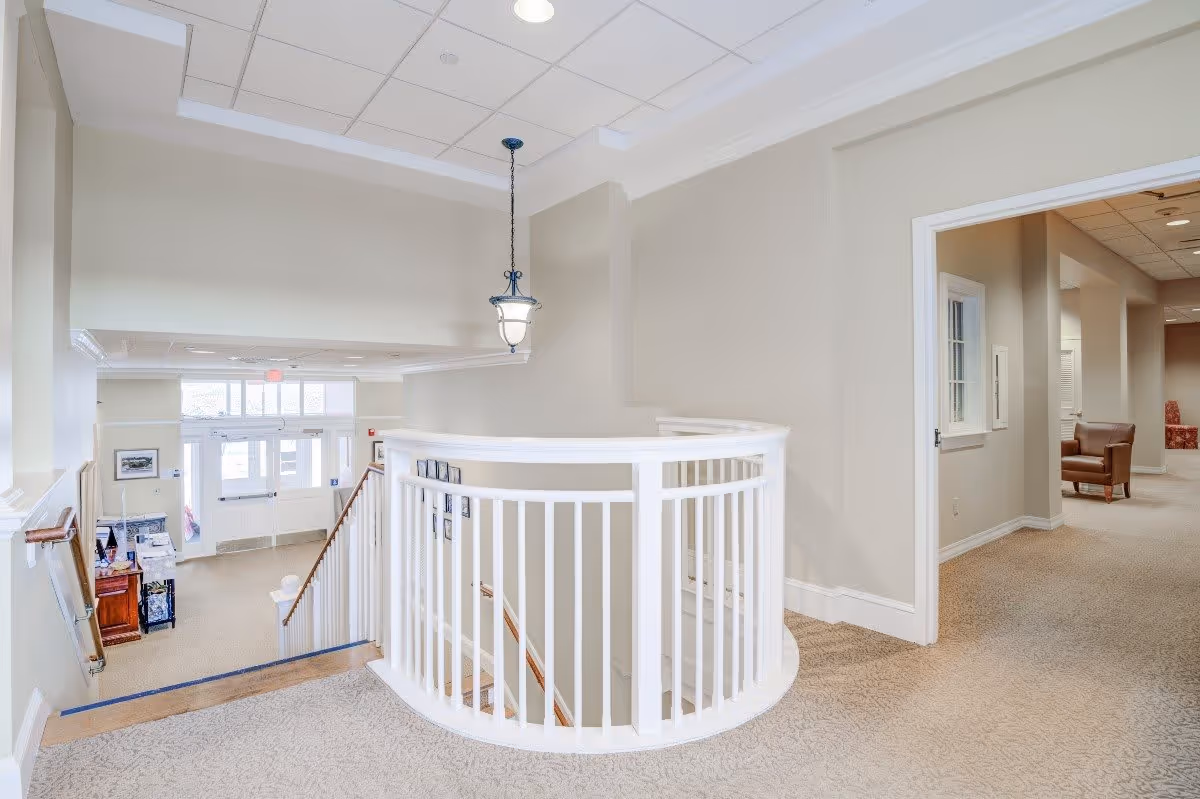 Interior view of a senior living facility showing a carpeted hallway with beige walls and white trim. A white wooden railing curves around a staircase leading downstairs. A hanging light fixture is suspended from the ceiling. In the background, there is a doorway leading to a room with chairs and further down a hallway. The entrance door with glass panels is visible at the bottom of the stairs.