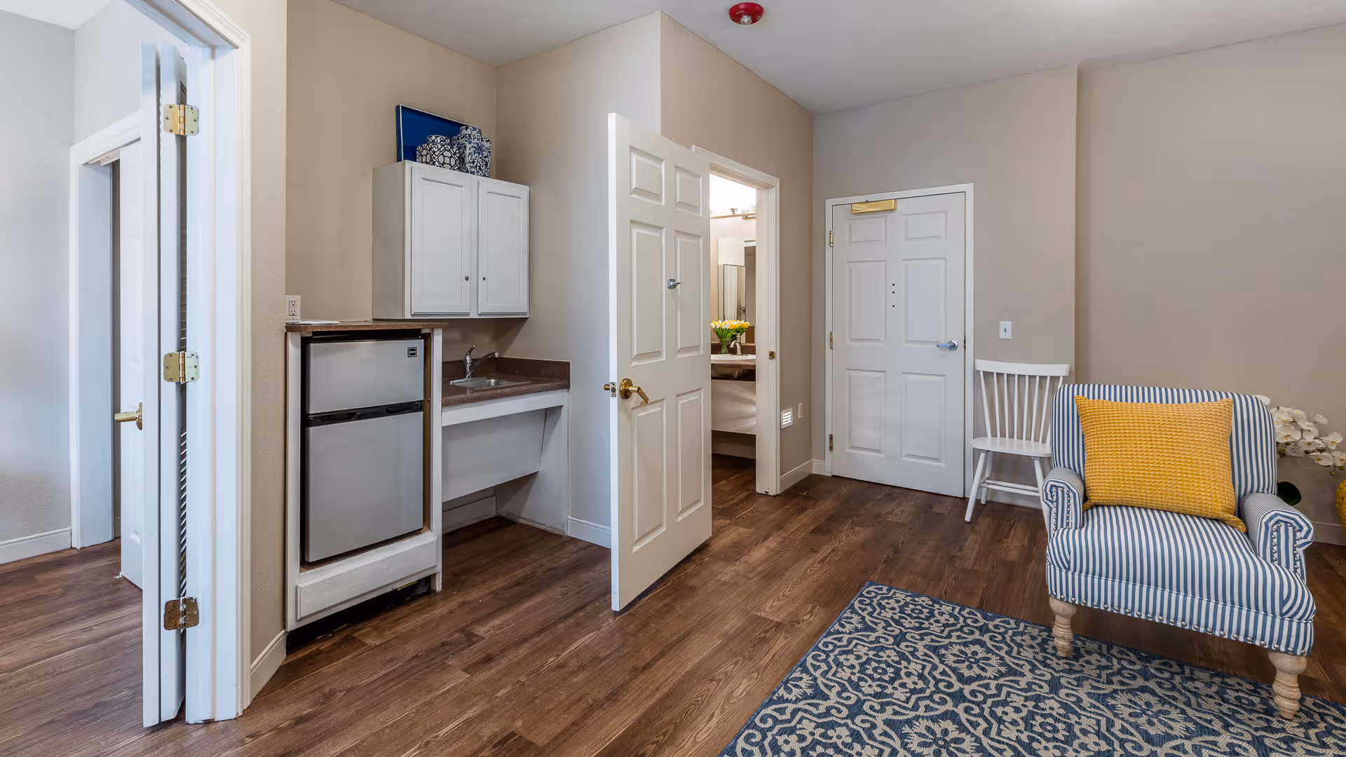 Bright assisted-living suite living area with a small kitchenette, striped armchair with a yellow pillow, wood floors and an open door to a bathroom/bedroom.