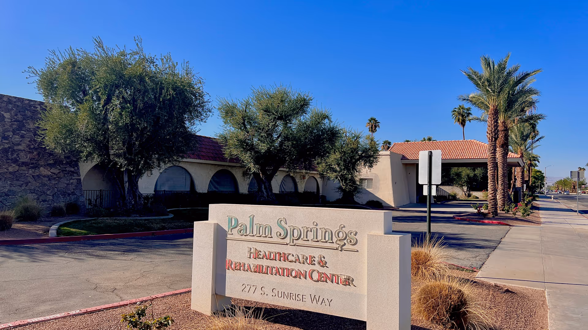 Exterior view of Palm Springs Healthcare & Rehabilitation Center with a large sign displaying the facility name and address. The building has a stone wall, arched windows, and a red-tiled roof. Palm trees and other greenery are visible along the sidewalk and driveway under a clear blue sky.
