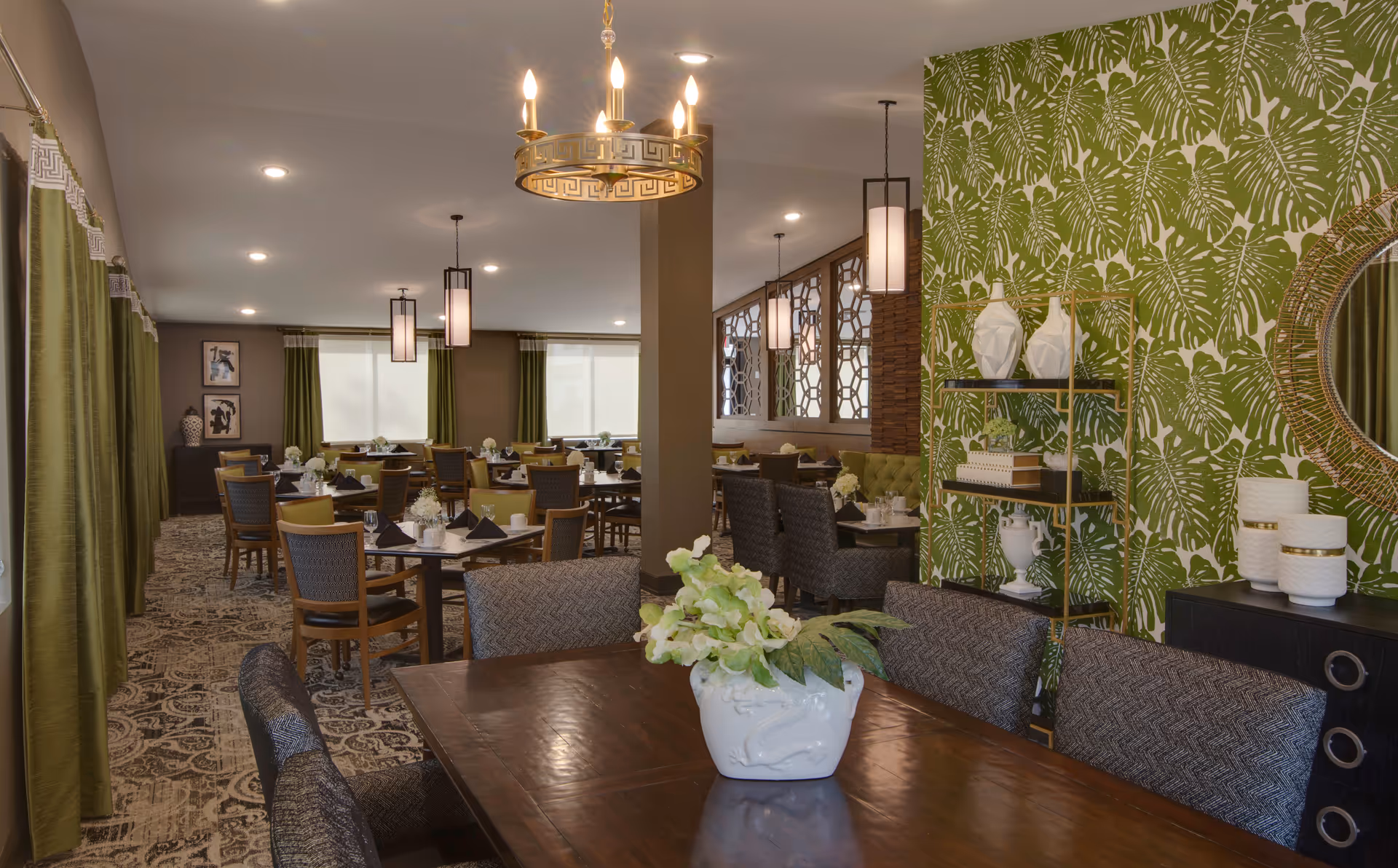 A well-decorated dining room with multiple tables and chairs arranged neatly. The room features green curtains, patterned carpet, and a green leafy wallpaper accent wall. There are modern light fixtures hanging from the ceiling and decorative items such as vases and a round mirror on a sideboard. A white vase with green and white flowers is placed on a wooden table in the foreground.