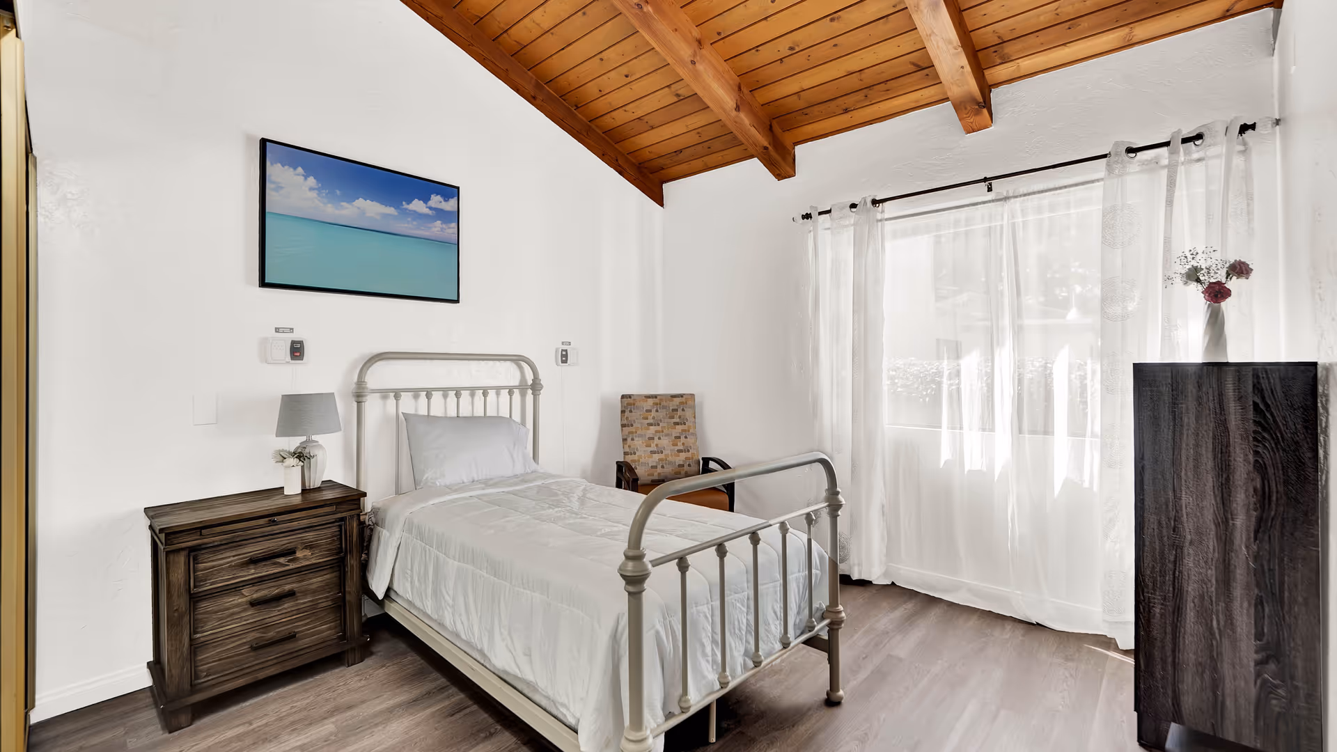 A bright bedroom with a single metal bed frame and white bedding. There is a wooden nightstand with a lamp and a small plant on it next to the bed. A patterned armchair is placed in the corner near a large window with sheer white curtains. The ceiling is wooden with exposed beams, and a framed picture of a serene ocean scene hangs on the wall above the bed.