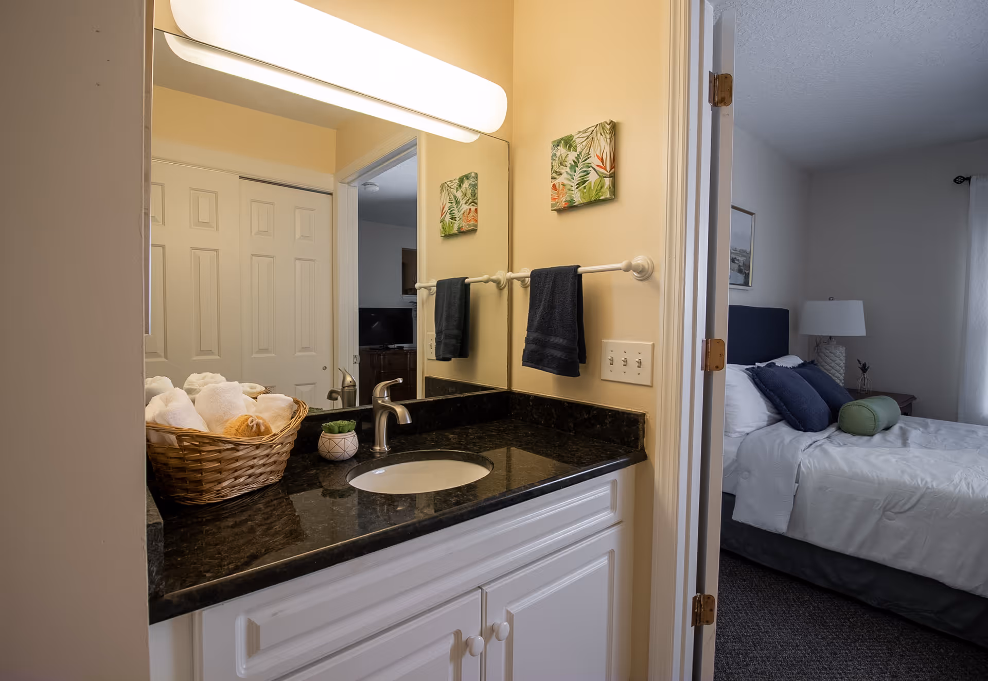 A bathroom vanity with a black countertop, white sink, and silver faucet. A basket with rolled white towels sits on the counter next to a small potted plant. A large mirror with a horizontal light fixture above reflects part of the room. To the right, an open door reveals a bedroom with a neatly made bed with white linens, blue and green pillows, a nightstand with a lamp, and a window with white curtains.