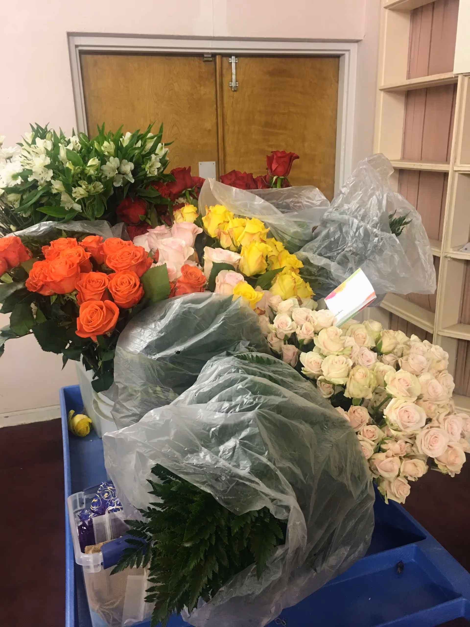A collection of various colorful flower bouquets including orange, yellow, pink, red roses, and white flowers arranged on a blue cart inside a room with wooden double doors and shelves in the background.