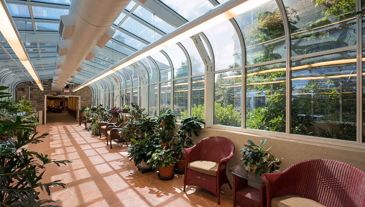 A bright indoor sunroom with large curved glass windows and ceiling, allowing natural light to flood the space. The floor is tiled with terracotta tiles, and there are several potted plants along the walls. Comfortable wicker chairs with cushions and small glass tables are arranged along the right side, creating a relaxing seating area. The far end leads to an interior hallway with stone walls.