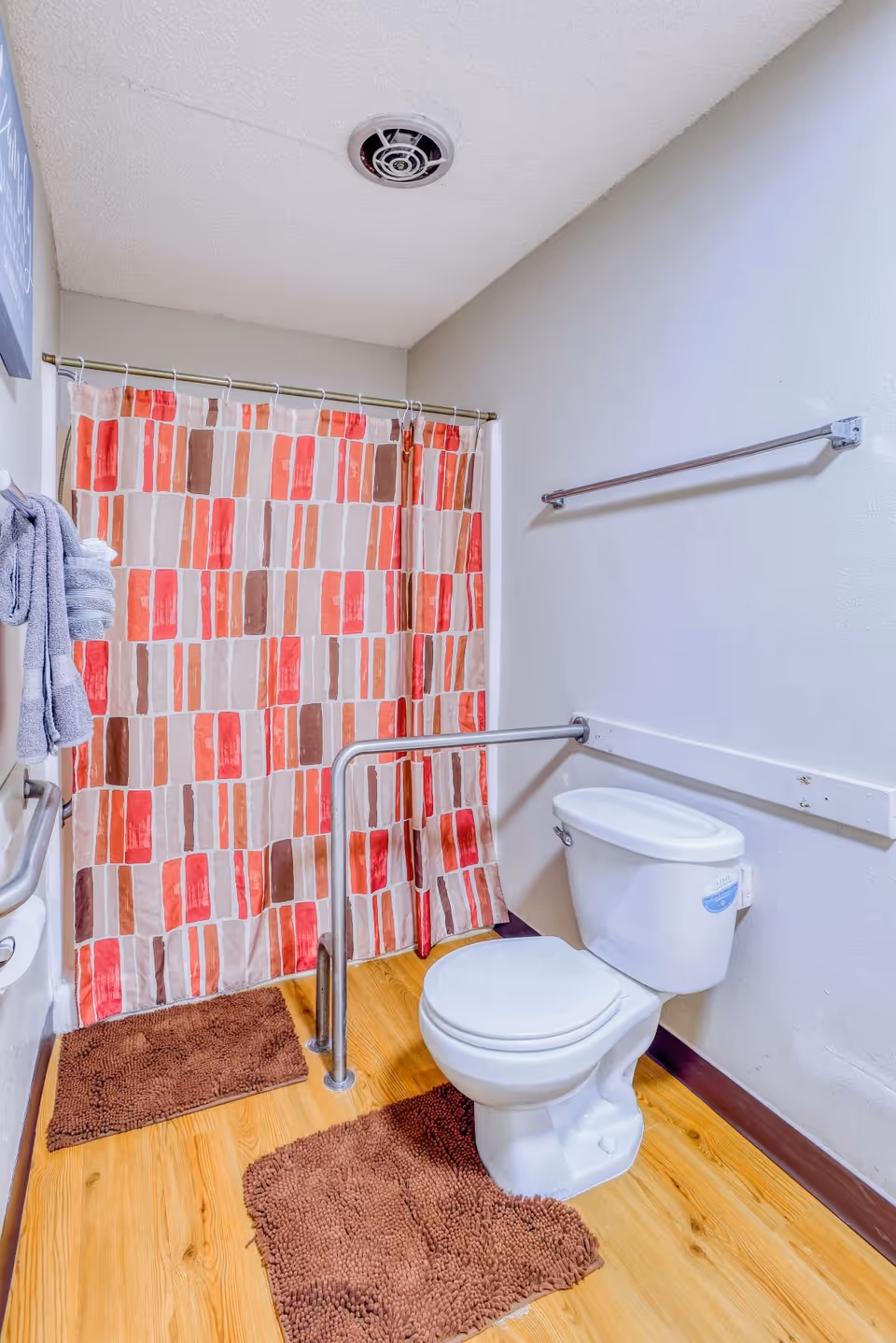 A bathroom with a white toilet, a safety grab bar around the toilet, a shower with a colorful red, orange, and beige patterned curtain, two brown bath mats on a wooden floor, a towel rack with a gray towel, and a ceiling vent.