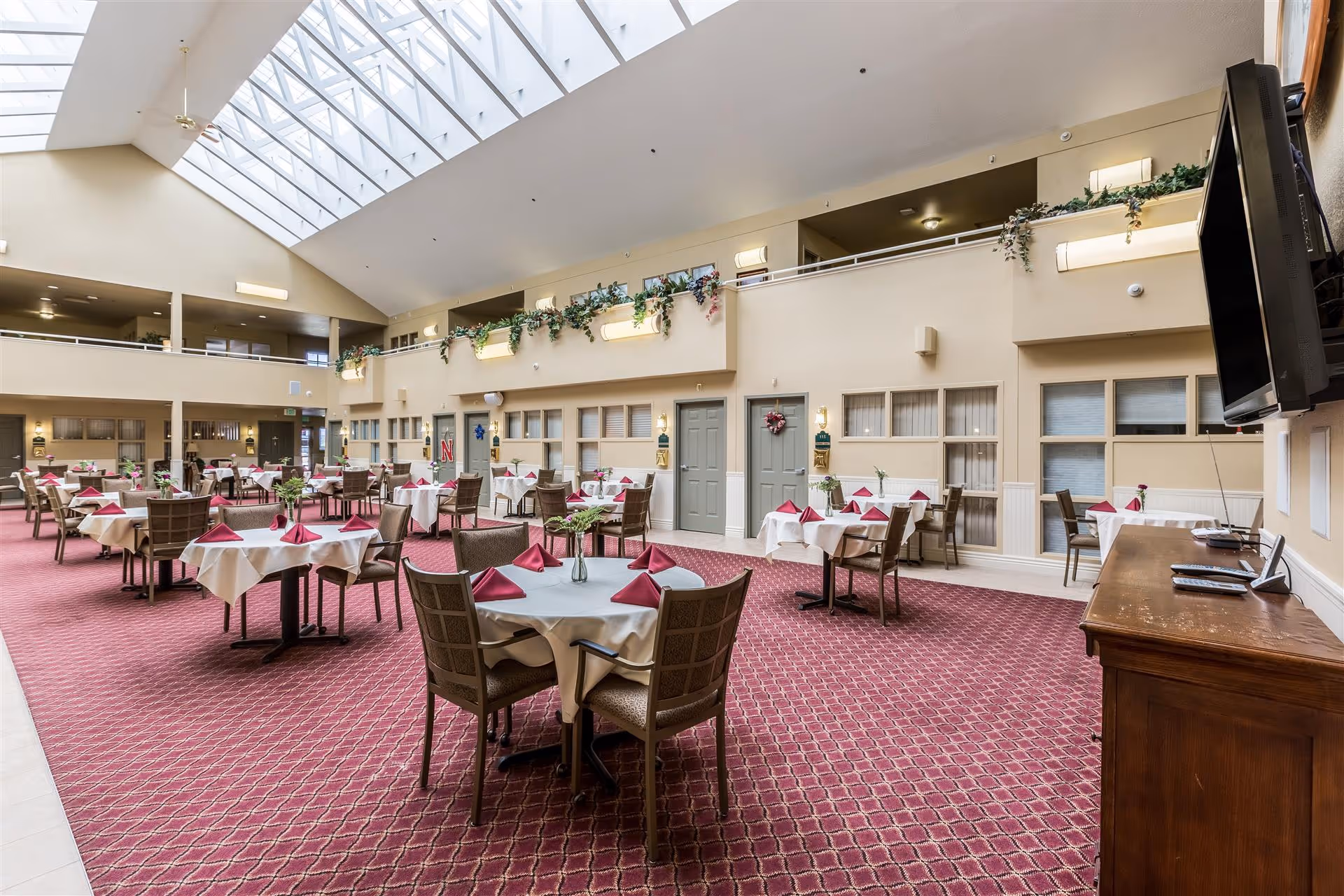 Spacious dining room with multiple round tables covered with white tablecloths and red napkins, surrounded by cushioned chairs. The room has a high ceiling with large skylights allowing natural light to fill the space. There are plants and greenery decorating the upper balcony area, and a large flat-screen TV mounted on the wall to the right.