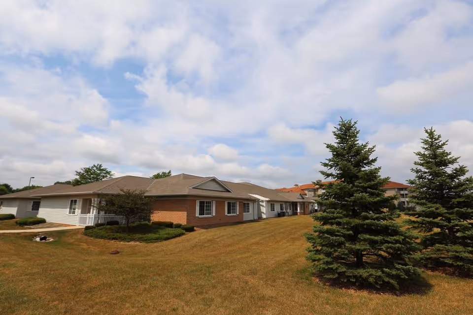 Exterior view of a single-story assisted living facility building with a combination of brick and white siding, surrounded by a grassy lawn and several evergreen trees under a partly cloudy sky.
