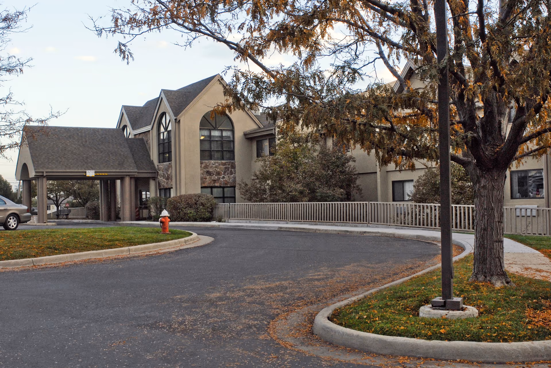 Exterior view of Life Care Center of Longmont showing a two-story building with large arched windows, a covered entrance, a curved driveway, a fire hydrant, and trees with autumn leaves.