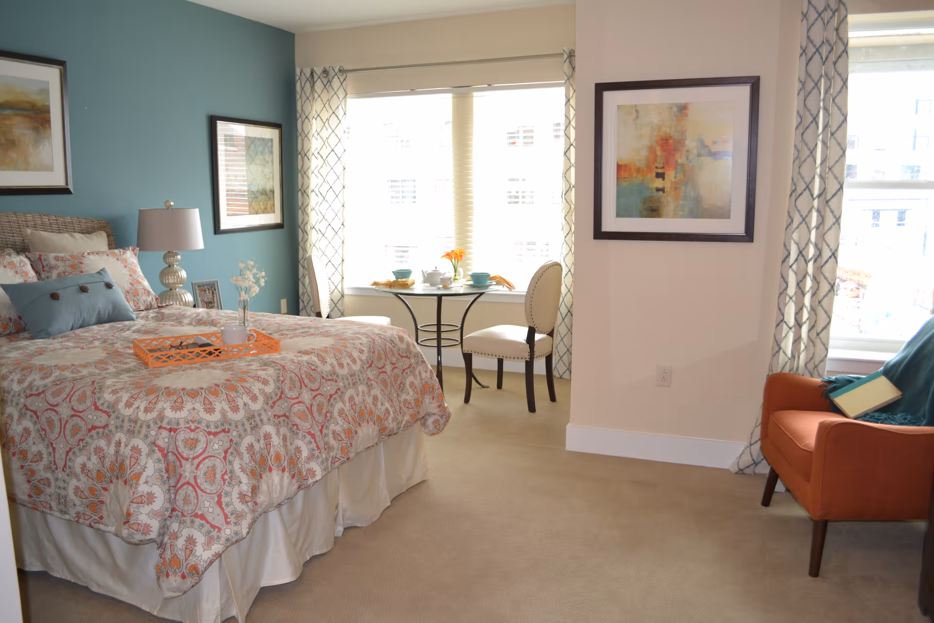 Bright, furnished bedroom with a patterned bed, bedside table, orange armchair and a small table and chairs by a large window.