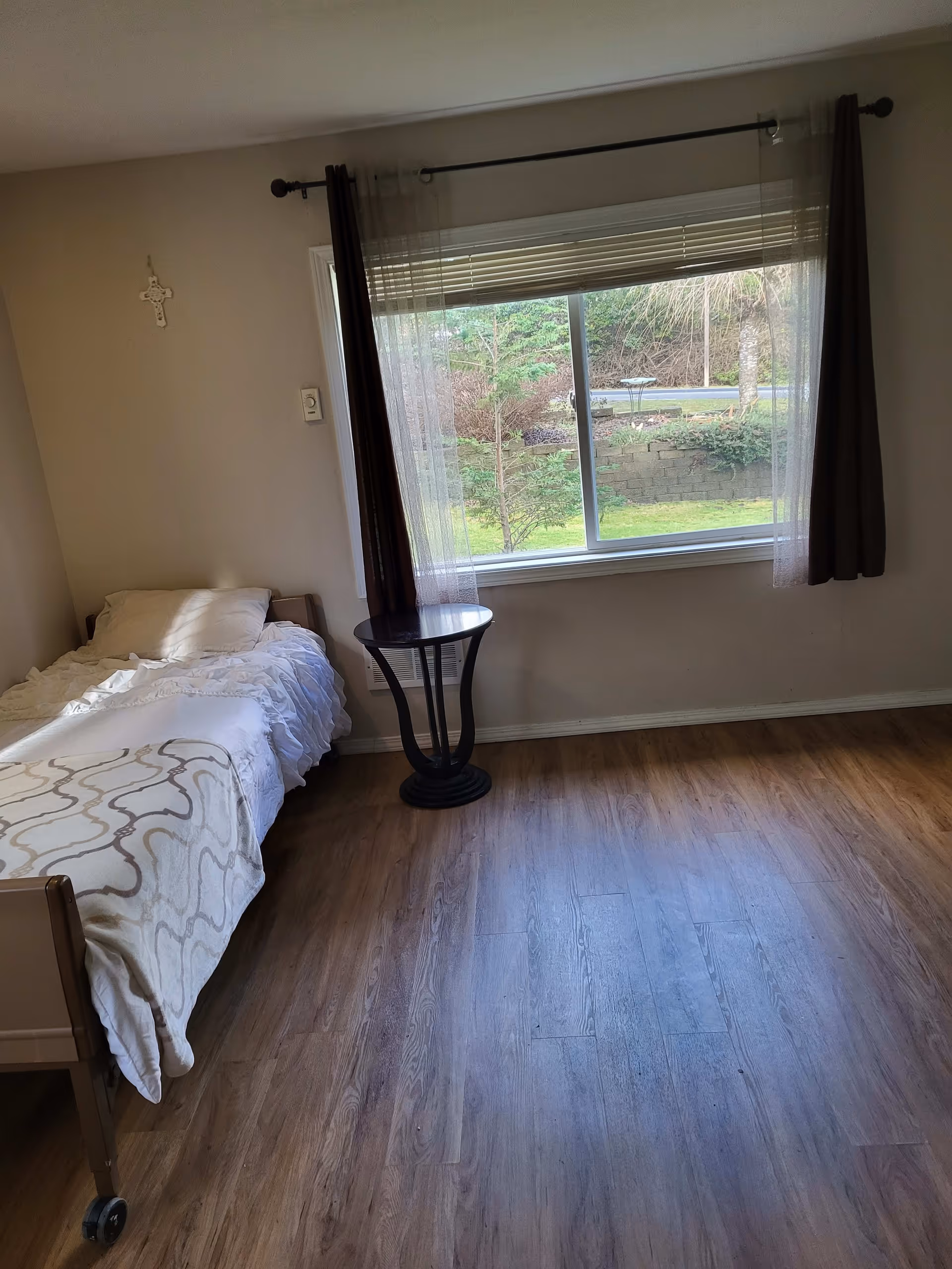 A simple bedroom with a single bed covered in white bedding and a patterned blanket. Next to the bed is a small round black side table. A large window with sheer and dark curtains lets in natural light and shows a view of greenery outside. The floor is wooden, and a decorative cross hangs on the wall above the bed.