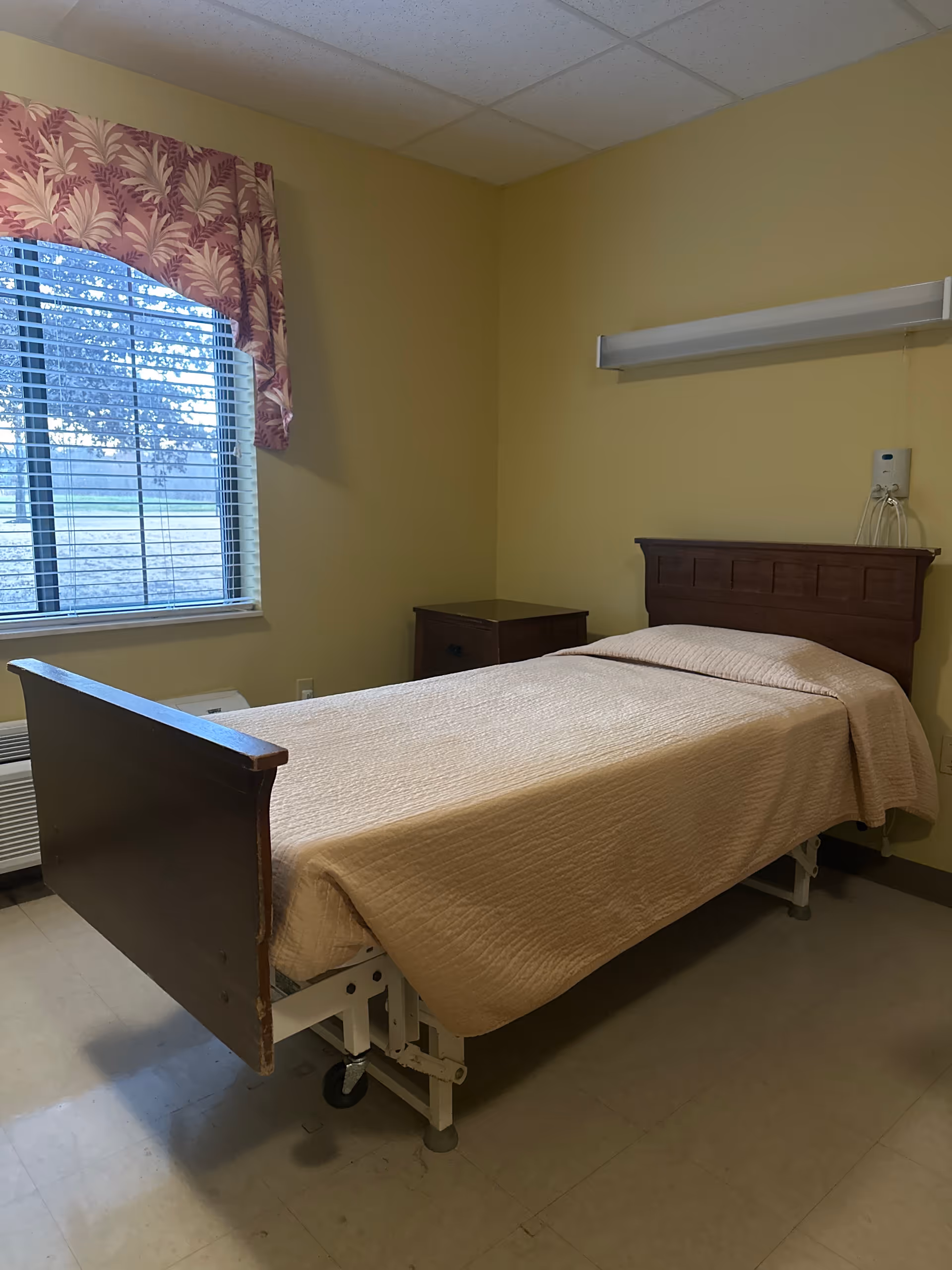 A simple room with a single hospital-style bed covered with a beige quilt. The bed has a wooden headboard and footboard and is on wheels. Next to the bed is a small wooden nightstand. A window with blinds and a floral valance is on the left wall, letting in natural light. The walls are painted light yellow, and the floor is tiled.