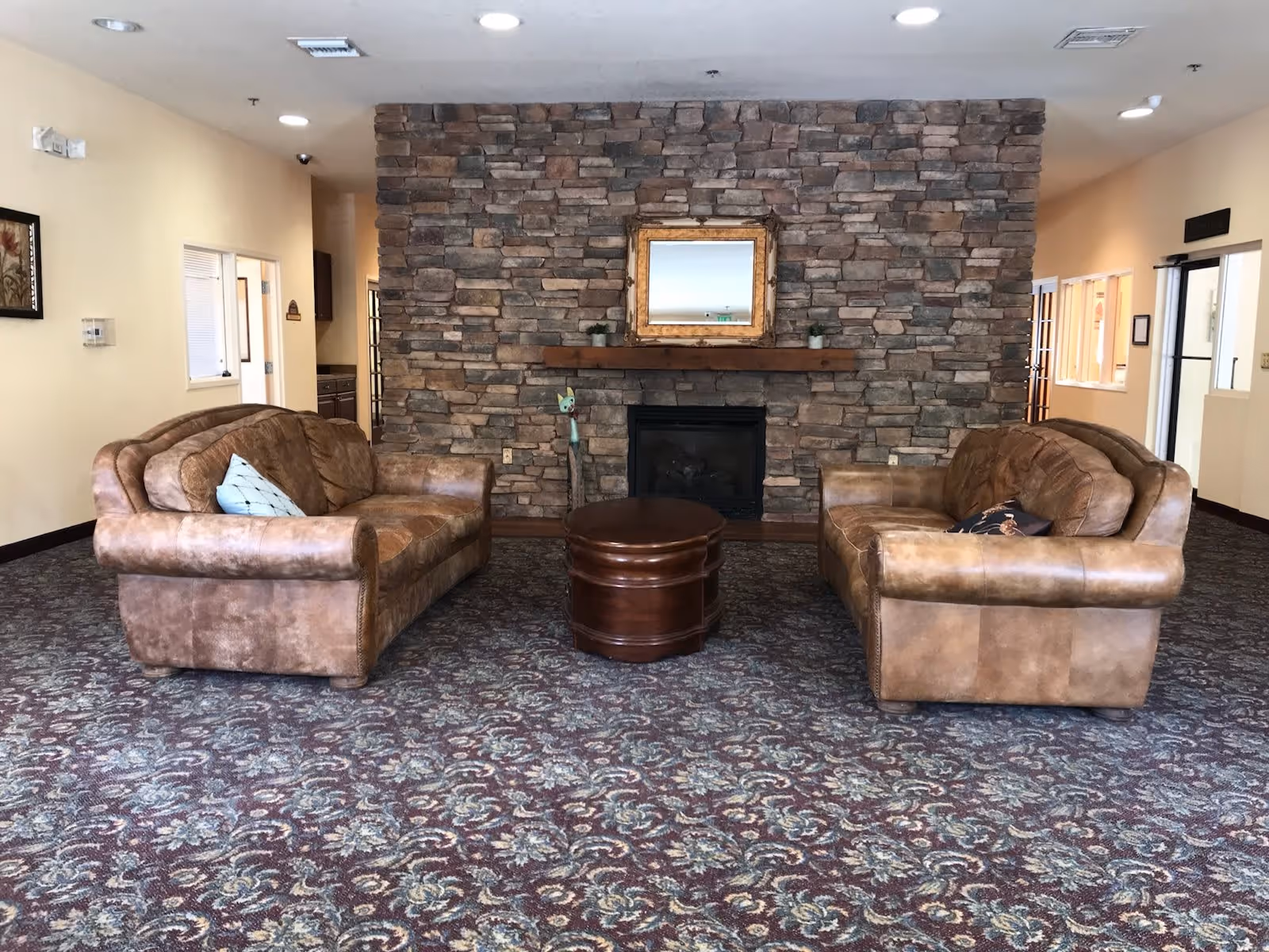 A cozy sitting area in a senior living facility with two brown leather sofas facing each other, a round wooden coffee table in the center, and a stone wall with a fireplace and a framed mirror above it. The room has patterned carpet flooring and beige walls with ceiling lights.