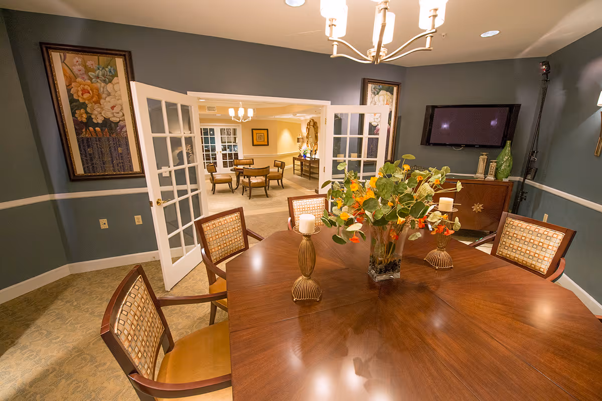 Well-lit dining/meeting room with a wooden table and chairs, a floral centerpiece, and French doors opening to another seating area.
