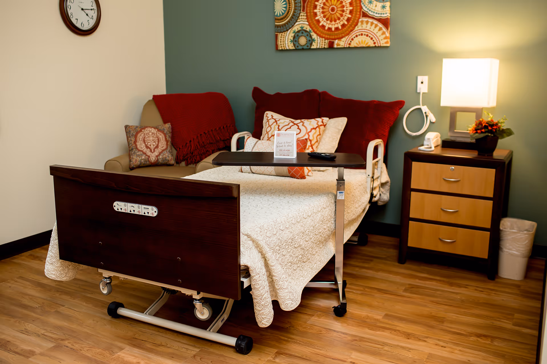 A cozy medical facility room featuring a hospital bed with a beige quilt, a dark wooden overbed table with a remote control and a small sign, a beige armchair with red and patterned pillows and a red throw blanket, a wooden nightstand with three drawers, a lamp, a small flower arrangement, a telephone, a wall clock, and a colorful circular patterned wall art above the bed.