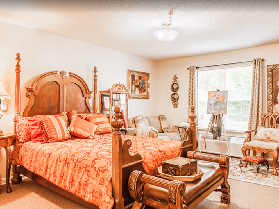 Ornately furnished bedroom with a wooden four-poster bed, red patterned bedding, seating area, and a window with curtains.