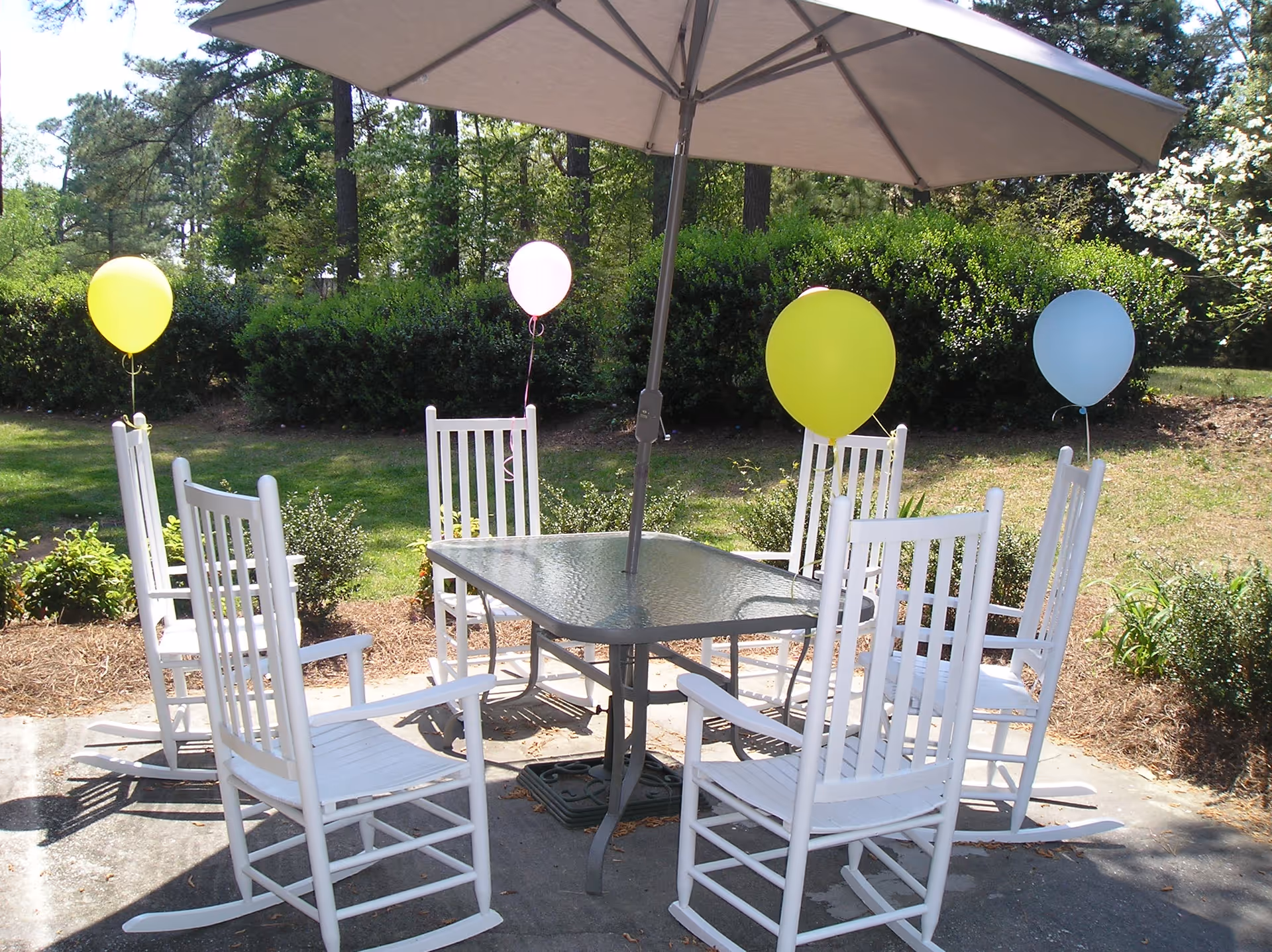 A backyard patio with six white rocking chairs around a glass table under a large umbrella, some chairs tied with colorful balloons.