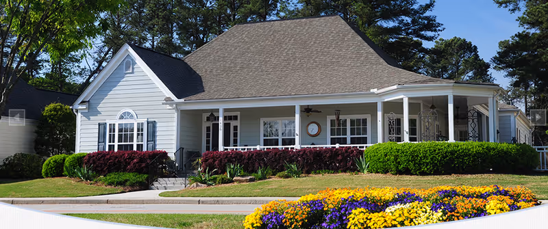 Exterior view of a single-story residential building with a large front porch, surrounded by well-maintained landscaping including colorful flower beds, green bushes, and trees in the background under a clear sky.