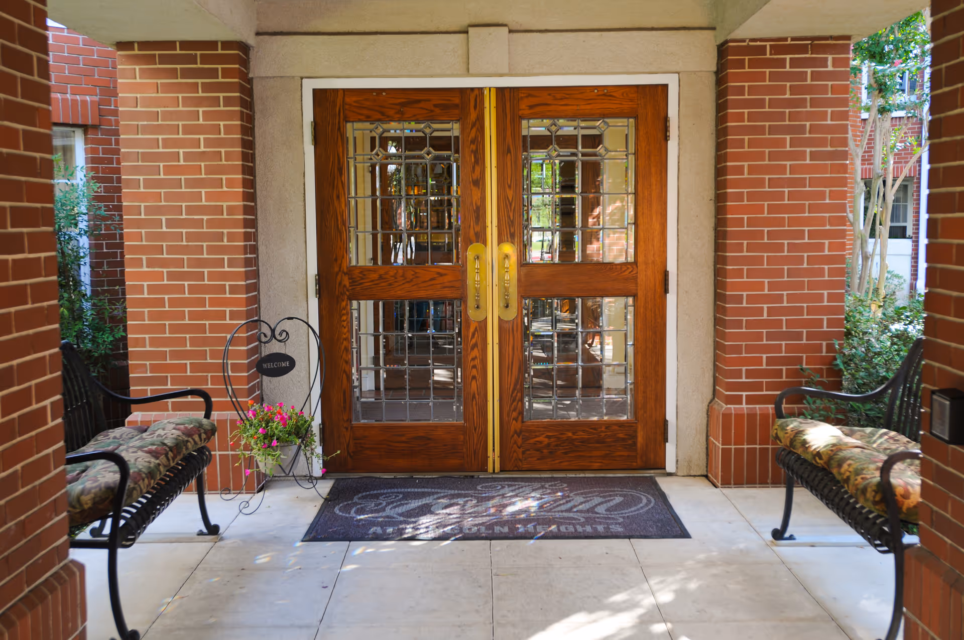 Entrance to The Forum at Lincoln Heights featuring double wooden doors with glass panels and brass handles, flanked by brick walls. Two cushioned benches are placed on either side of the entrance, and a small decorative stand with a welcome sign and flowers is on the left. A welcome mat with the facility name is on the floor in front of the doors.