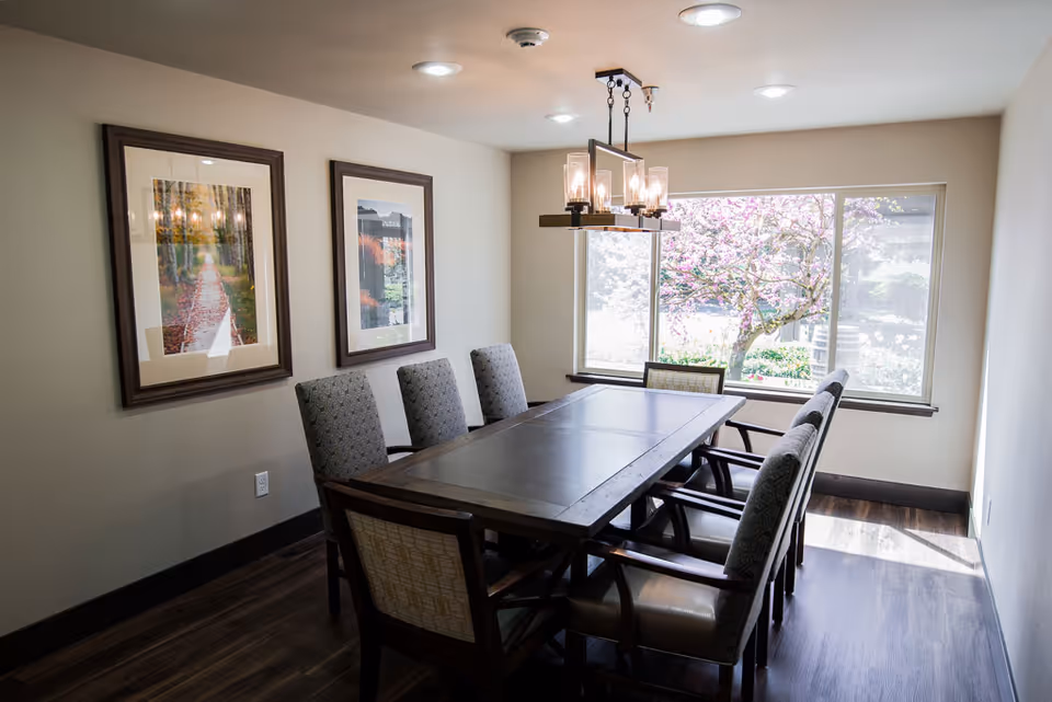 A dining room with a long rectangular wooden table surrounded by eight upholstered chairs. Two framed pictures hang on the left wall, and a large window on the right side shows a blooming tree outside. A modern chandelier with multiple light bulbs hangs above the table, and the floor is dark wood.