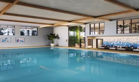 Indoor swimming pool with lounge chairs, potted plants, and large windows in a residential facility.