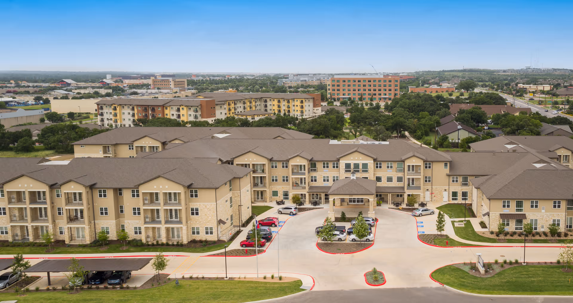 Aerial view of The Enclave at Round Rock Senior Living facility showing multiple beige multi-story buildings with brown roofs arranged around a central driveway and parking area. The surrounding area includes greenery, trees, and other buildings in the distance under a clear sky.