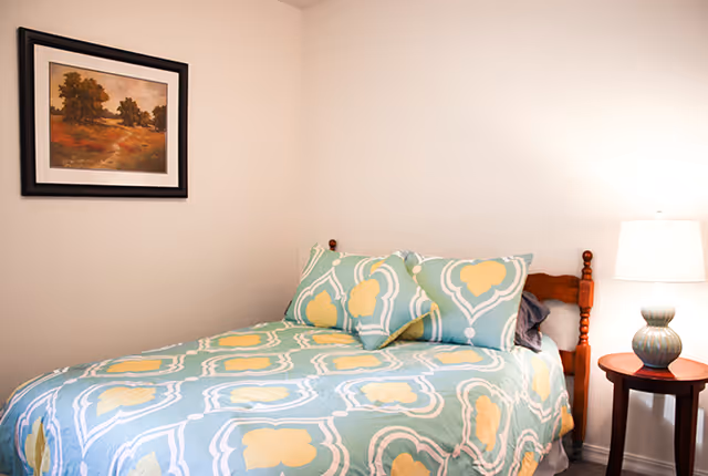 A bedroom with a wooden bed frame, a bed covered with a blue, yellow, and white patterned comforter and pillows. There is a wooden nightstand with a lamp on it to the right of the bed, and a framed landscape painting hanging on the wall above the bed.