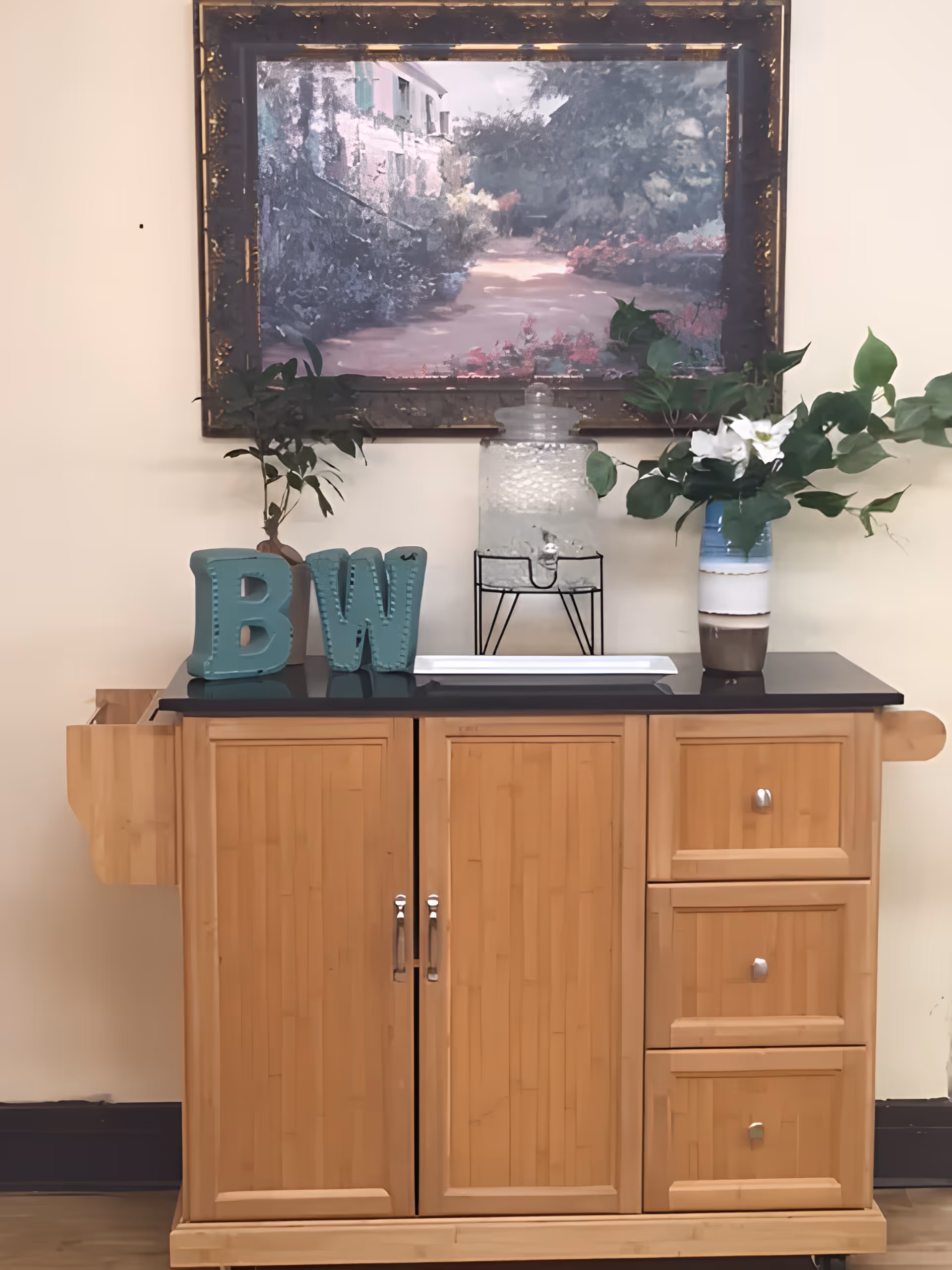 A wooden cabinet with a black countertop, decorated with a glass water dispenser filled with ice, a vase with green leaves and white flowers, and teal decorative letters 'B' and 'W'. Above the cabinet hangs a framed painting of a garden pathway with flowers and greenery.