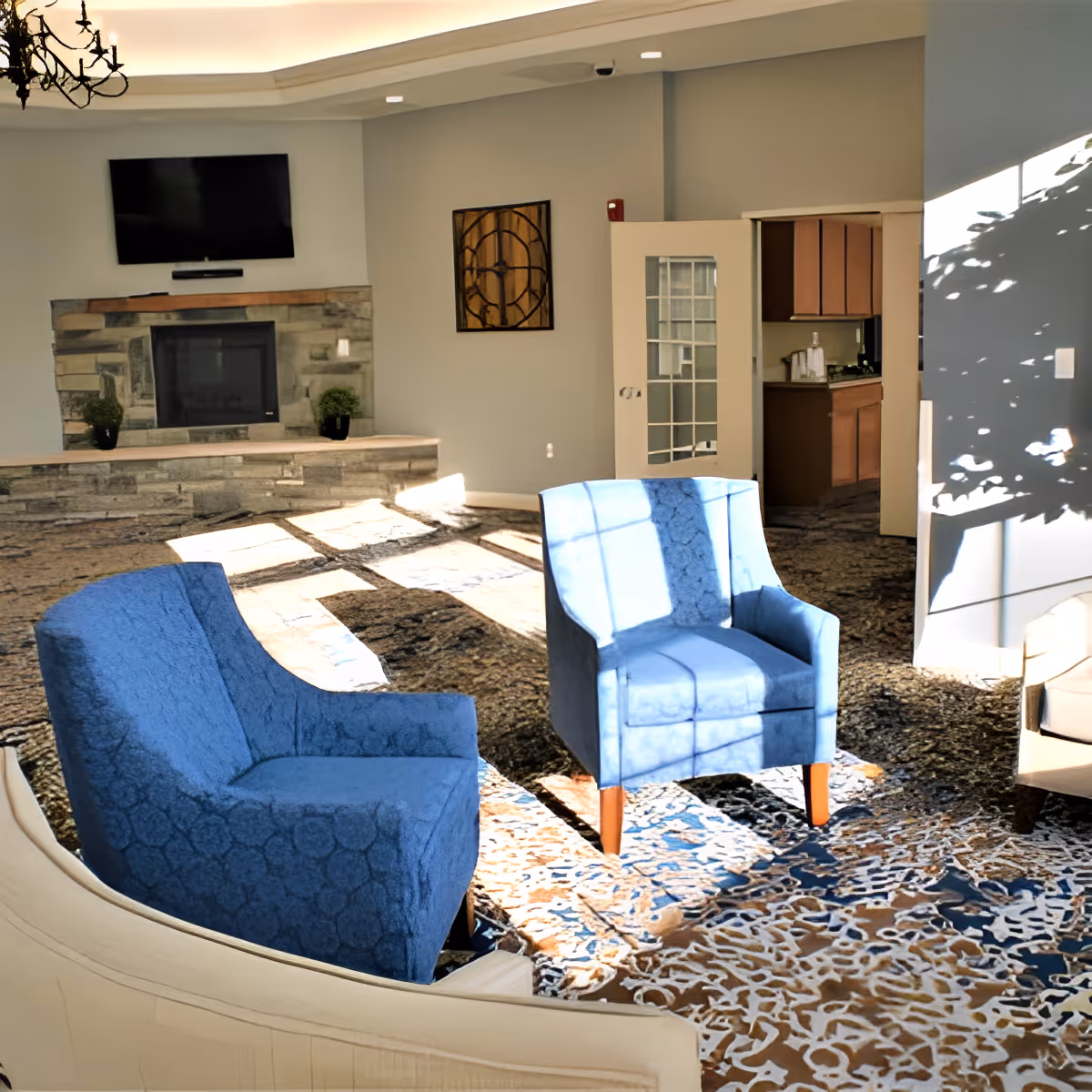 A bright living room area with two blue upholstered armchairs and a beige sofa arranged on a patterned carpet. The room features a stone fireplace with a mounted flat-screen TV above it, a decorative wall clock, and a door leading to a small kitchenette. Sunlight streams through windows, casting shadows on the furniture and floor.