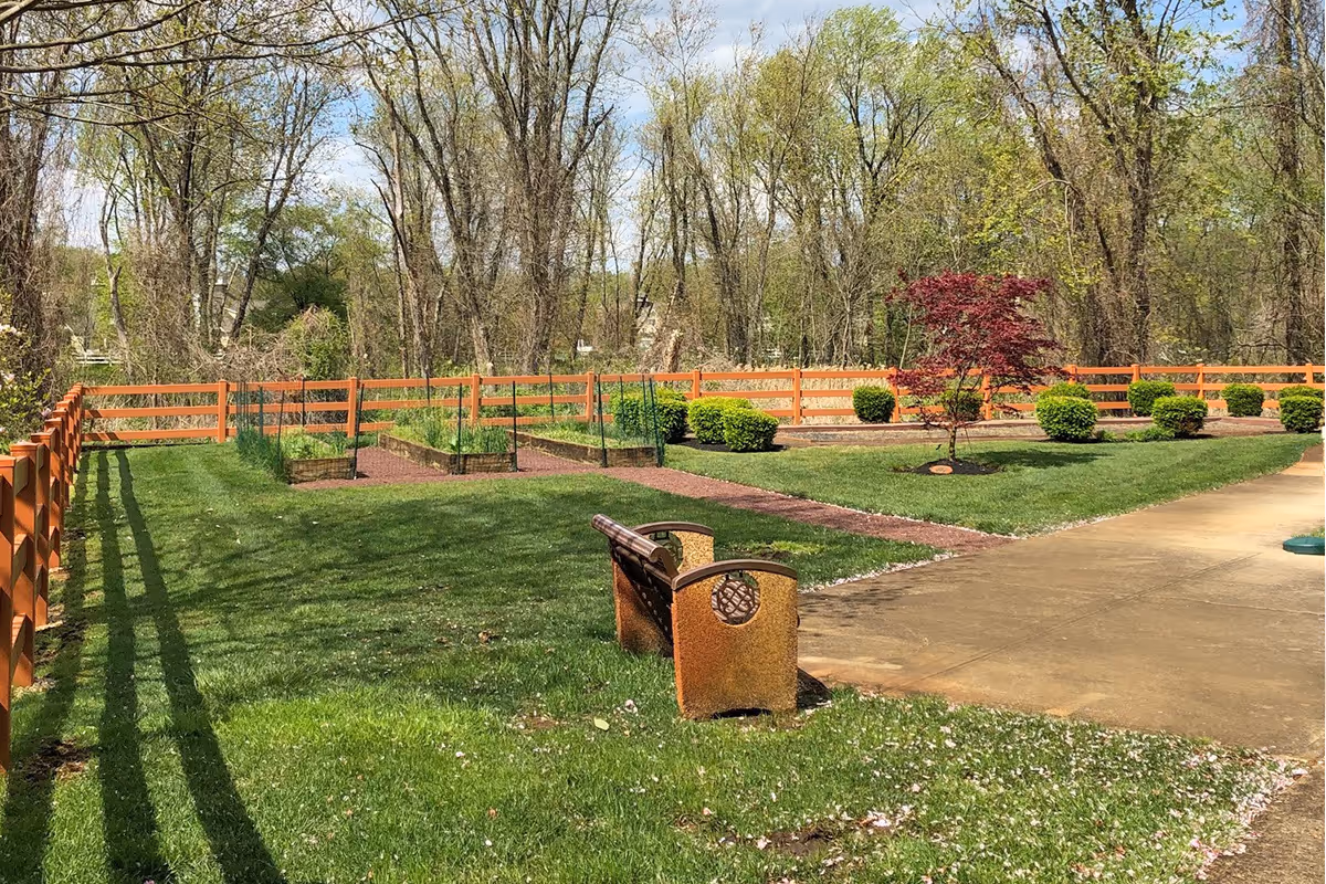 A peaceful outdoor garden area with a wooden fence, green grass, several small bushes, a red-leafed tree, and raised garden beds. There is a wooden bench facing the garden beds and a paved walkway on the right side. Tall trees are visible in the background under a partly cloudy sky.