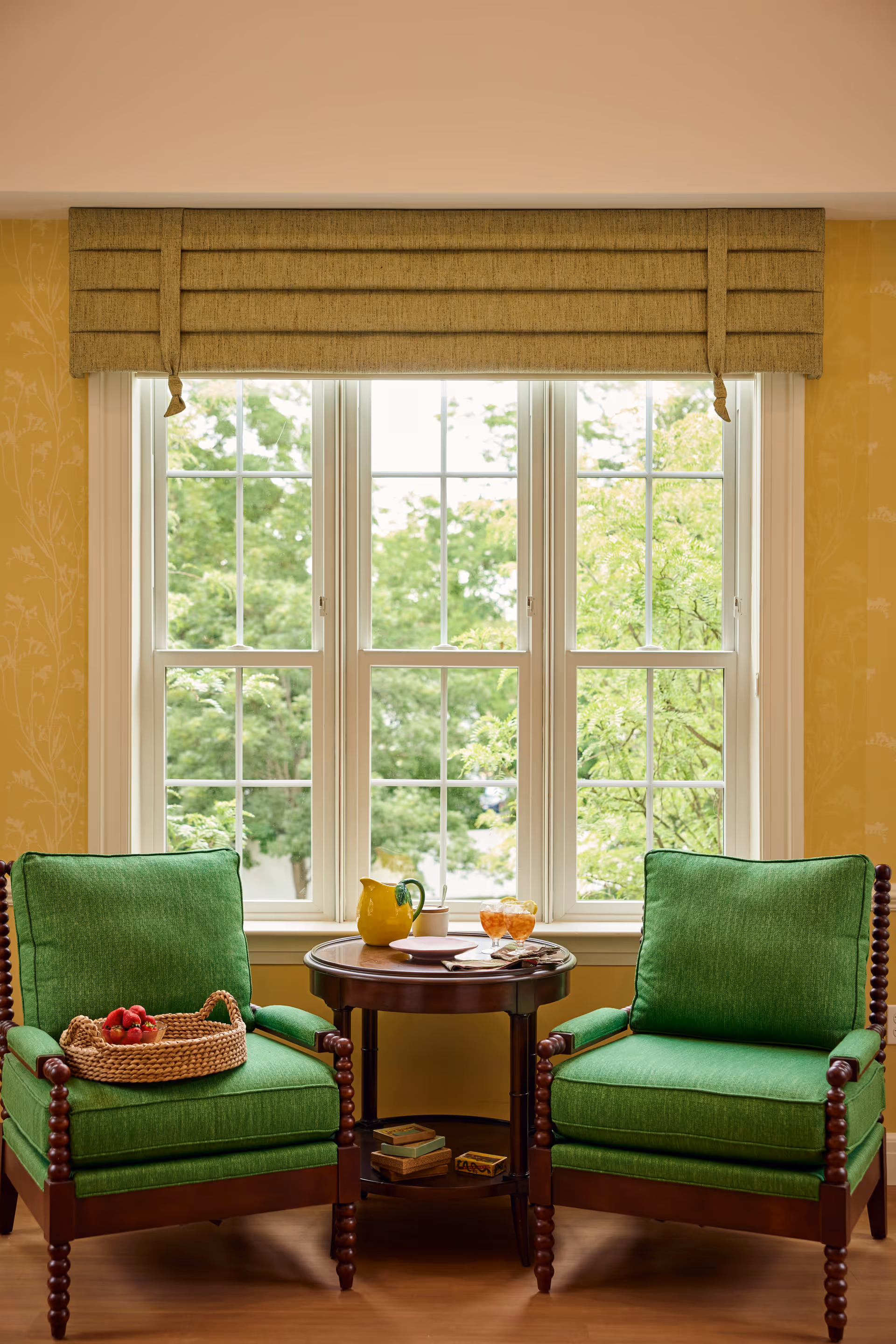 Two green cushioned wooden armchairs with a small round wooden table between them, placed in front of a large window with white frames and a beige valance. The table holds a yellow pitcher, a cup, a bowl of orange snacks, and some reading material. A basket with red apples is on one of the chairs. Outside the window, green trees are visible.