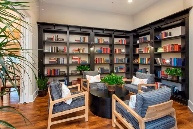 A cozy reading room with dark wooden bookshelves filled with books lining the walls. Four cushioned armchairs with wooden frames are arranged around a round black coffee table with a potted plant on it. The room has wooden flooring and is well-lit with ceiling lights.