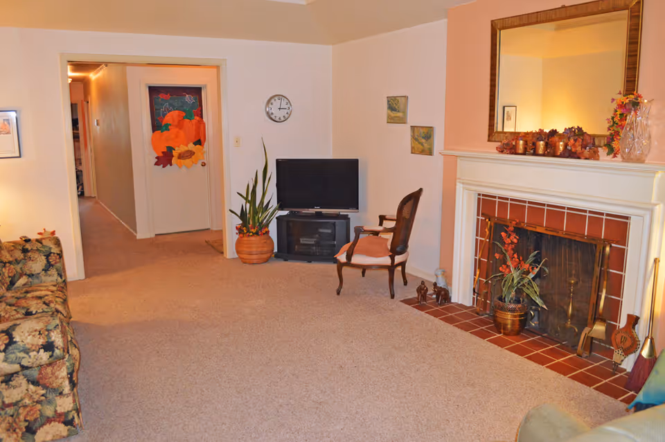 A cozy living room with beige carpet, a floral patterned sofa on the left, a wooden chair with a cushion near a TV on a stand, and a fireplace decorated with autumn-themed items and a large mirror above it. A hallway and a door decorated with a pumpkin and sunflower fall-themed decoration are visible in the background.