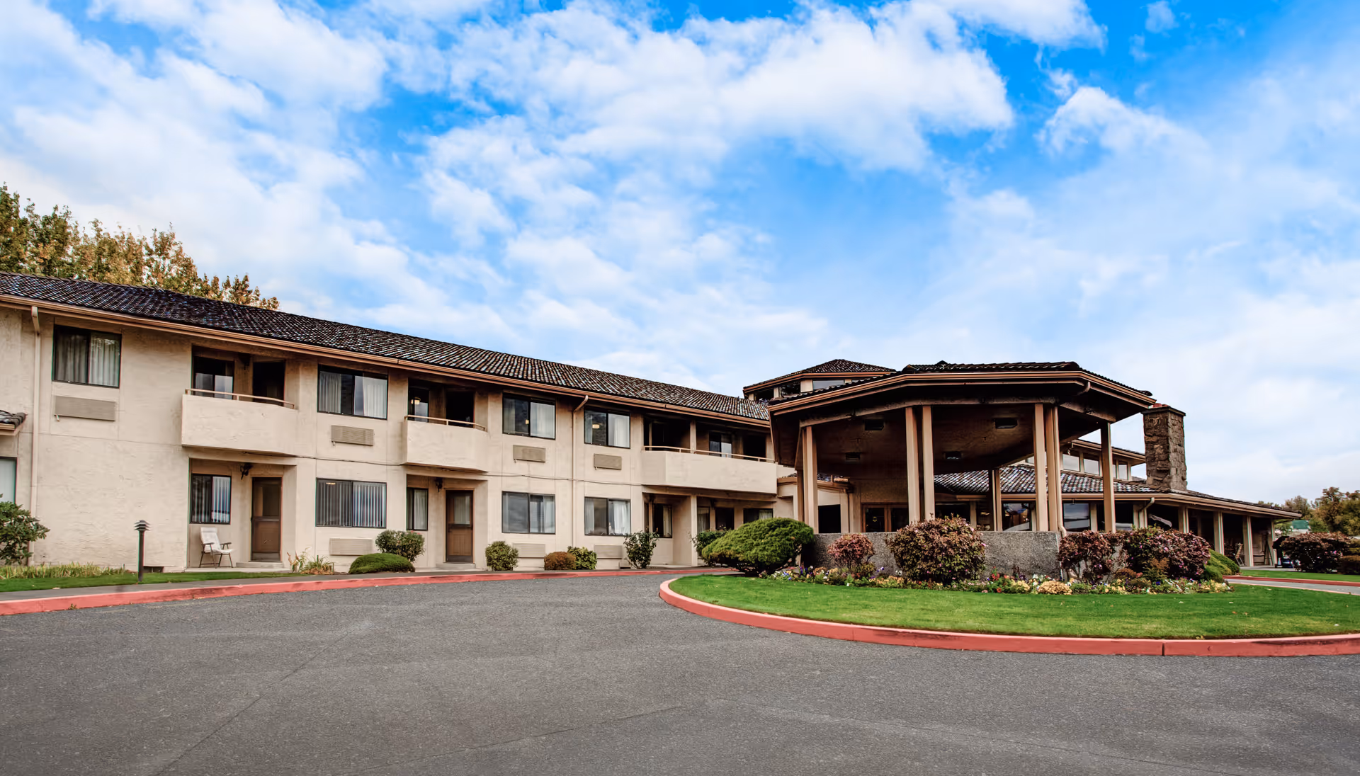 Exterior view of a two-story senior living facility building with beige walls and a tiled roof. The building has multiple windows and balconies. There is a covered entrance area supported by columns, surrounded by landscaped bushes and a green lawn. The sky is partly cloudy.