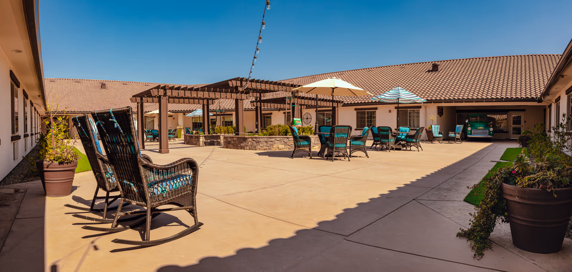 Outdoor courtyard area of El Rio Memory Care Community with wicker rocking chairs and tables with umbrellas arranged on a spacious concrete patio. The courtyard is surrounded by single-story buildings with tiled roofs and has a pergola structure in the center. Potted plants are placed along the edges of the patio under a clear blue sky.