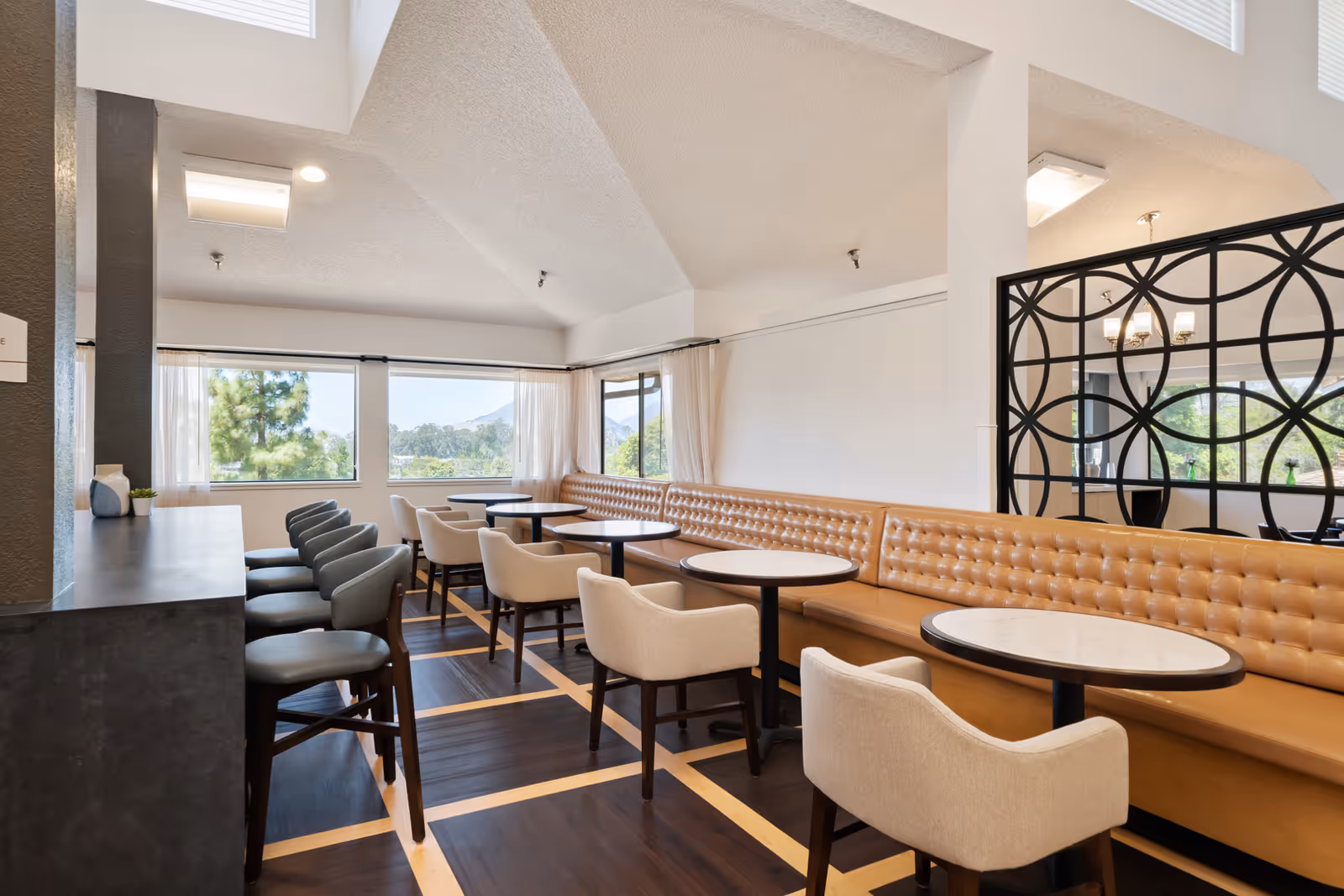 A bright dining area with multiple round tables and cushioned chairs. There is a long tan tufted bench along one wall, and several individual chairs on the opposite side of the tables. Large windows let in natural light and offer views of trees and mountains outside. The room features a dark wood floor with light wood inlays and a decorative black partition with circular patterns.