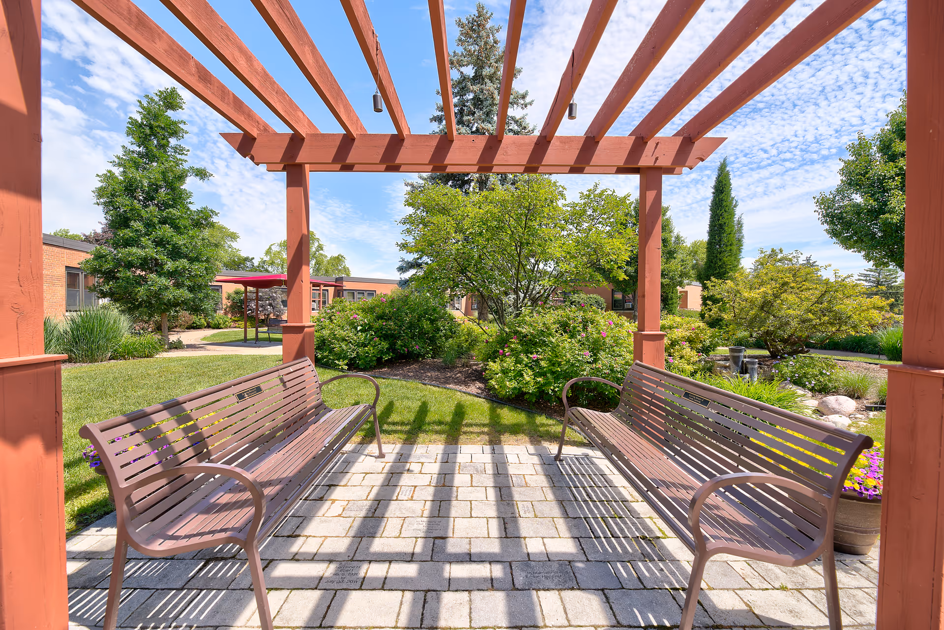 Outdoor seating area with two metal benches under a wooden pergola surrounded by green trees, bushes, and a well-maintained garden on a sunny day.