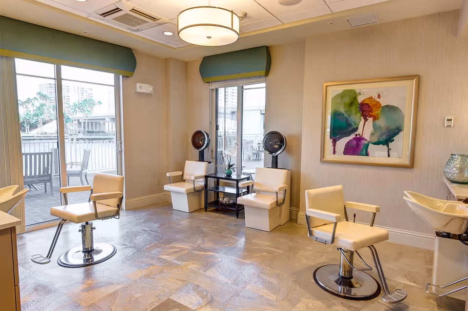 Interior of a salon area in a senior living facility with beige salon chairs, two hair drying stations, a framed colorful abstract painting on the wall, large windows with green valances, and a door leading to an outdoor patio with chairs and tables.