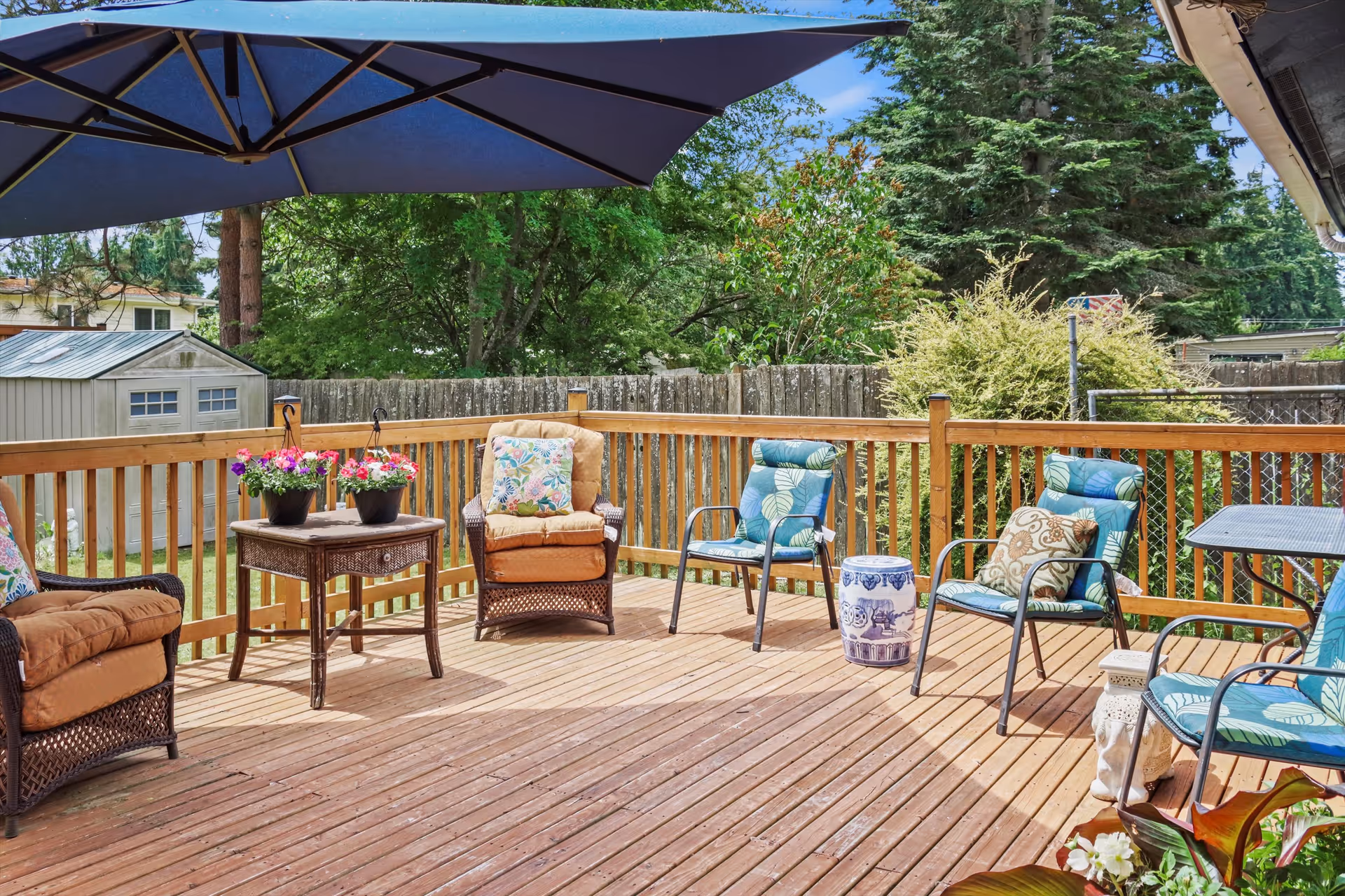 A wooden outdoor deck with several cushioned chairs, a small table with flower pots, a large blue patio umbrella, and a wooden fence surrounding the area. Trees and a shed are visible in the background.