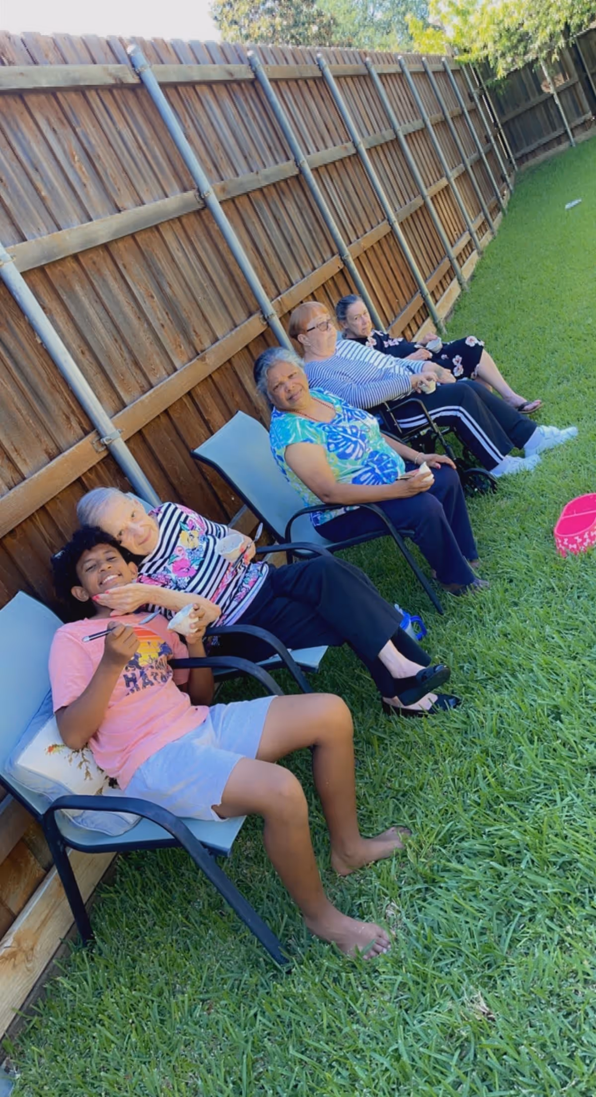 Five people sitting in chairs along a wooden fence in a grassy backyard. Four elderly women and one young boy are seated, enjoying a sunny day outdoors. The boy is smiling and holding a spoon, while the elderly women appear relaxed and content.