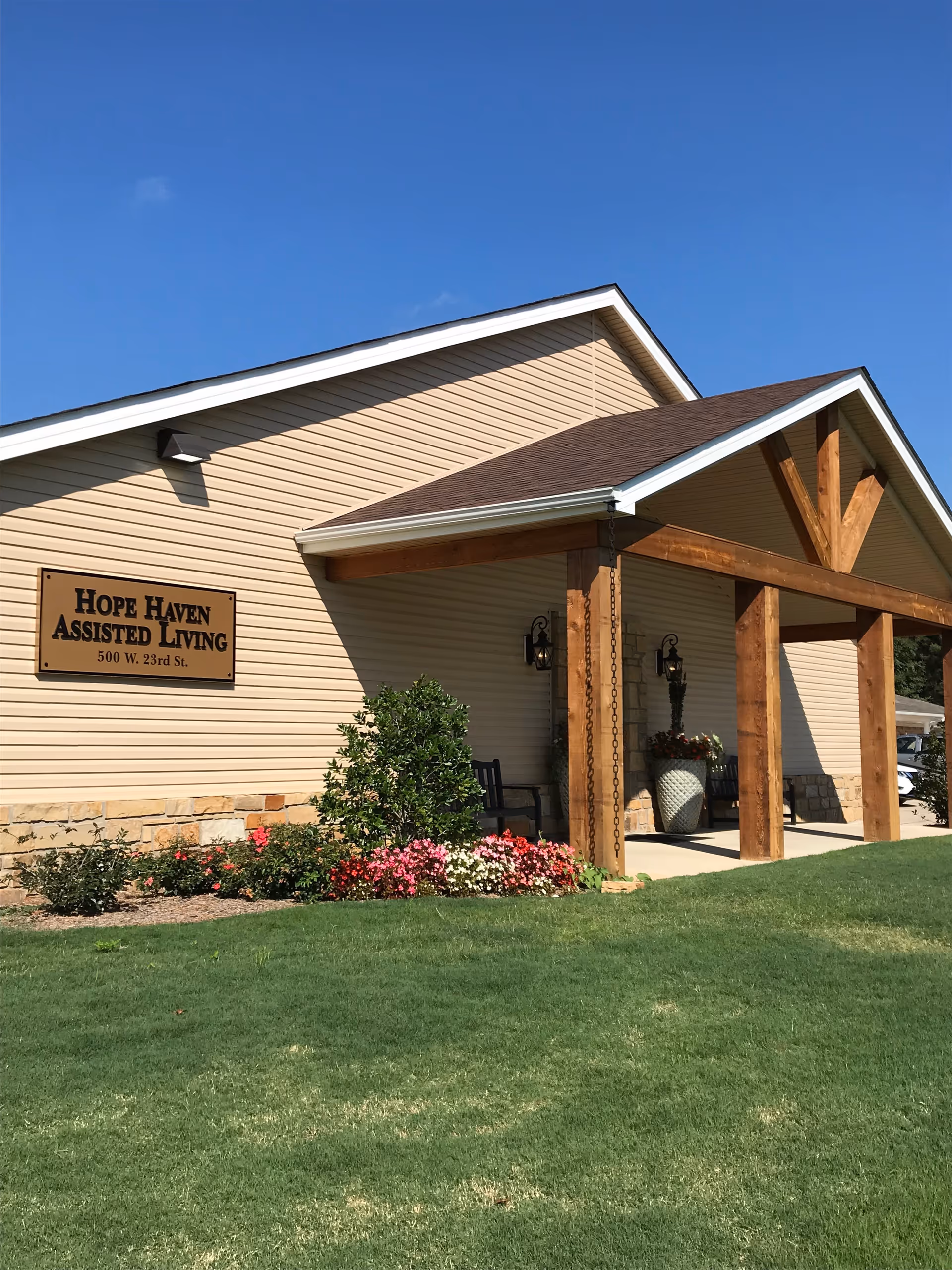 Exterior view of Hope Haven Assisted Living Community building with beige siding, a brown roof, wooden beams supporting a covered entrance, green lawn, and colorful flower beds under a clear blue sky.