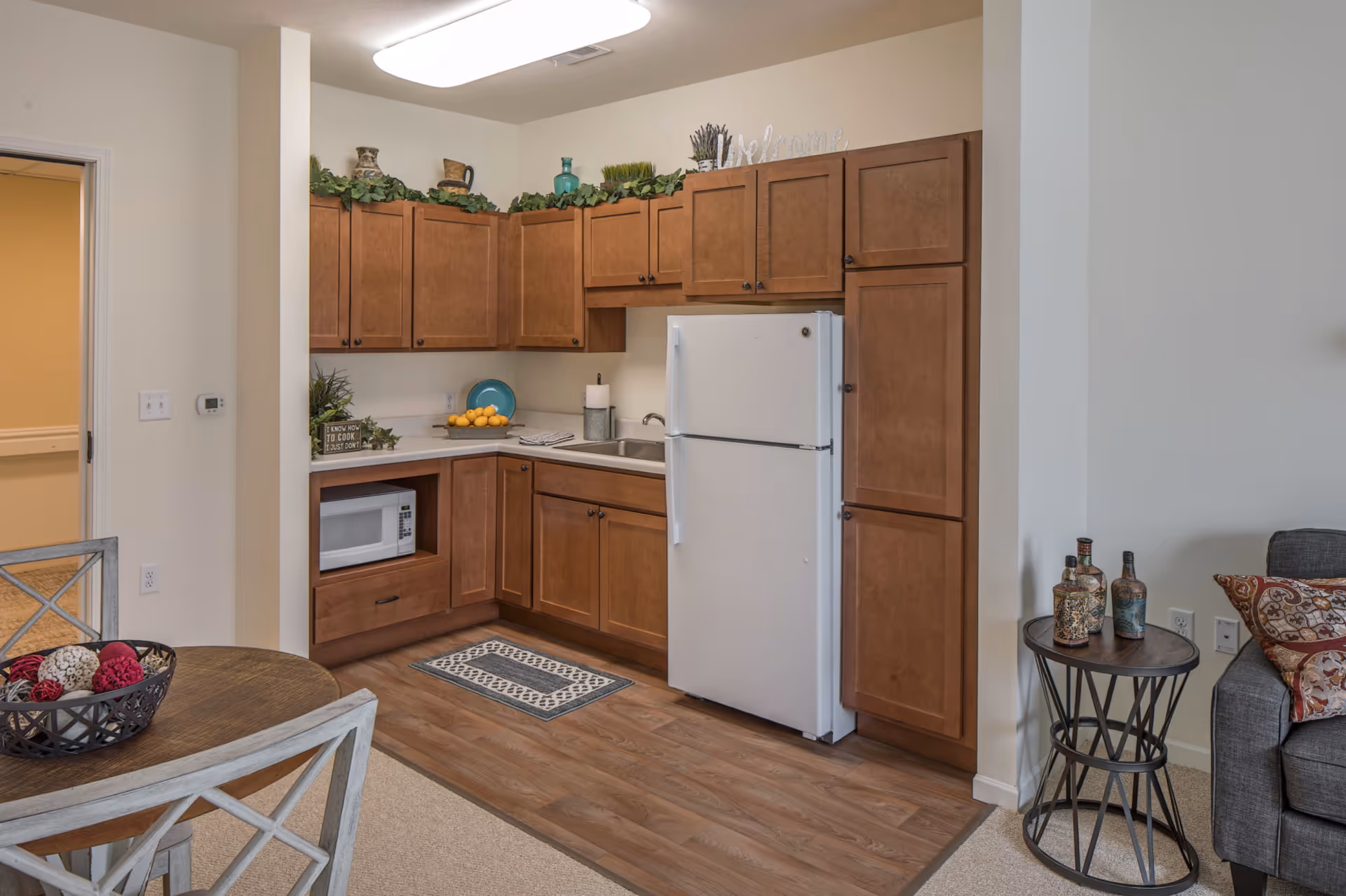 A small kitchen area with wooden cabinets, a white refrigerator, a microwave, and a sink. The countertop has a bowl of lemons, a blue plate, and a paper towel holder. Above the cabinets are decorative items including plants and vases. To the left is a round wooden dining table with chairs, and to the right is a small round side table with decorative bottles next to a gray sofa with patterned pillows.