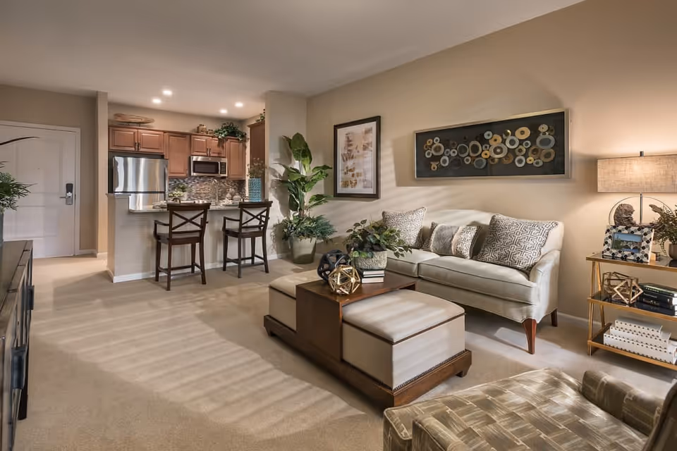 Open-plan living room and kitchen with a sofa, ottoman coffee table, bar stools, and decorative artwork.