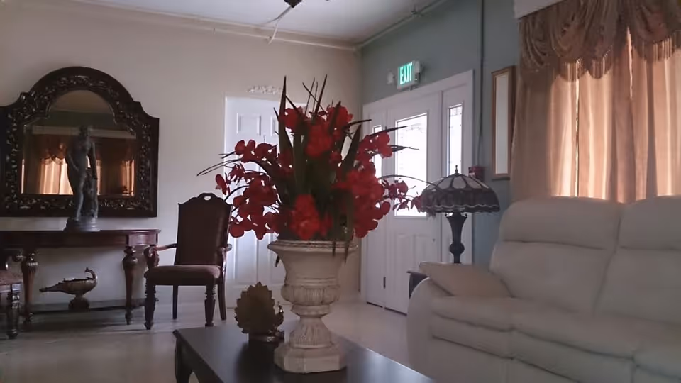 A living room area with a beige sofa on the right, a coffee table with a large vase of red flowers in the center, and a wooden chair and table with a decorative statue and mirror on the left. There is a door with glass panels and an exit sign above it in the background, and a window with beige curtains on the right.