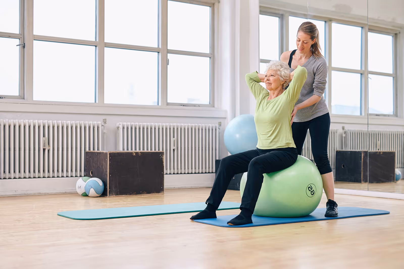 An elderly woman in a green shirt and black pants sits on a green exercise ball while a younger woman in athletic wear assists her with a stretch in a bright exercise room with large windows and wooden floors.