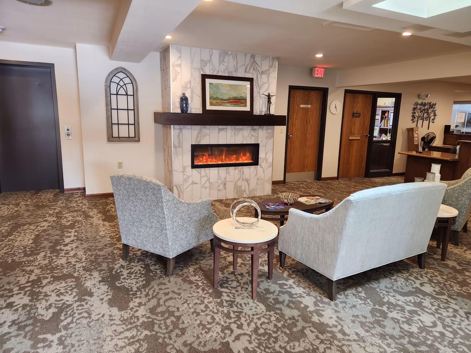 A cozy seating area in a senior living facility with two upholstered armchairs and a loveseat arranged around a small round table and a rectangular coffee table. The room features a modern electric fireplace with a marble surround and a wooden mantel, above which hangs a framed landscape painting. The carpet has a floral pattern, and there are wooden doors and a reception desk visible in the background.