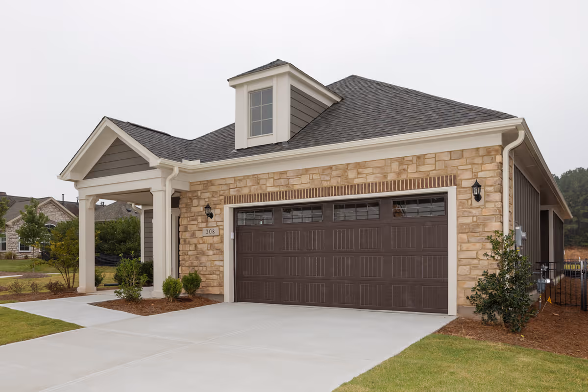Exterior view of a single-story residential building with a stone facade and a dark brown garage door. The building has a small covered porch supported by white columns and a dormer window on the roof. There is a concrete driveway and some landscaping with small bushes and grass.