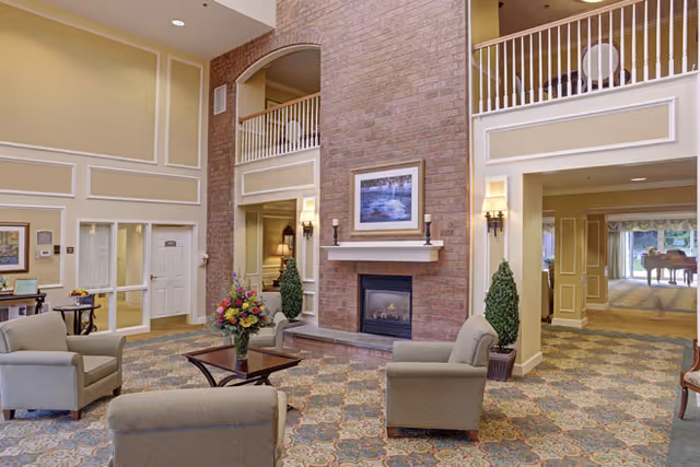 A spacious senior living facility common area with a high ceiling and a large brick fireplace in the center. The room is furnished with several cushioned armchairs arranged around a wooden coffee table with a flower arrangement. There are wall sconces on either side of the fireplace, framed artwork above it, and decorative plants flanking the hearth. The carpet has a patterned design, and there is a balcony railing visible on the upper level. In the background, there is a hallway leading to another room with a grand piano.