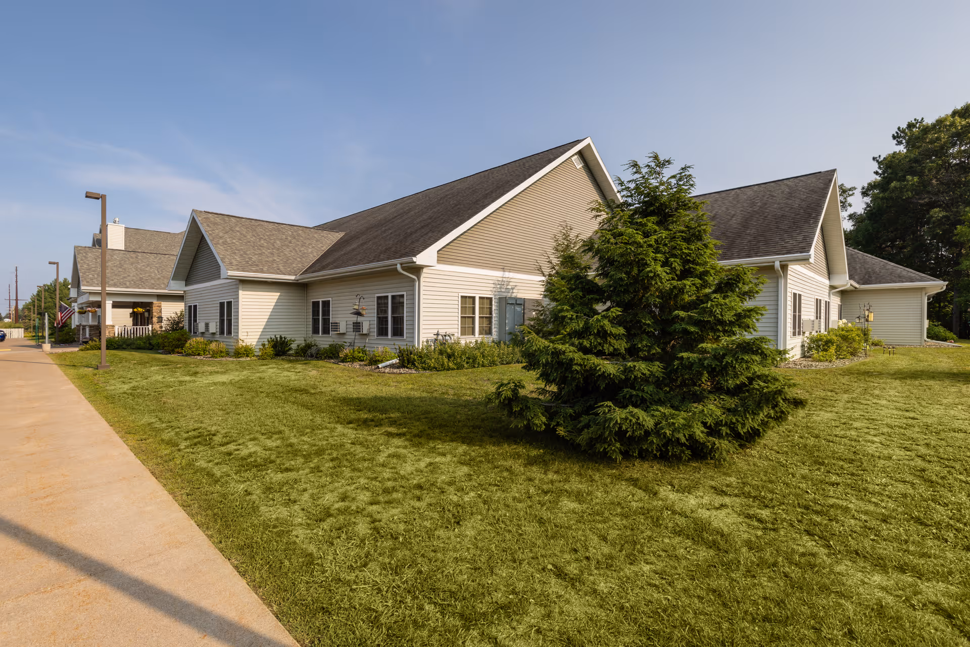 Exterior view of a single-story senior living facility building with beige siding and a gray shingled roof, surrounded by green grass and landscaping, under a clear blue sky.